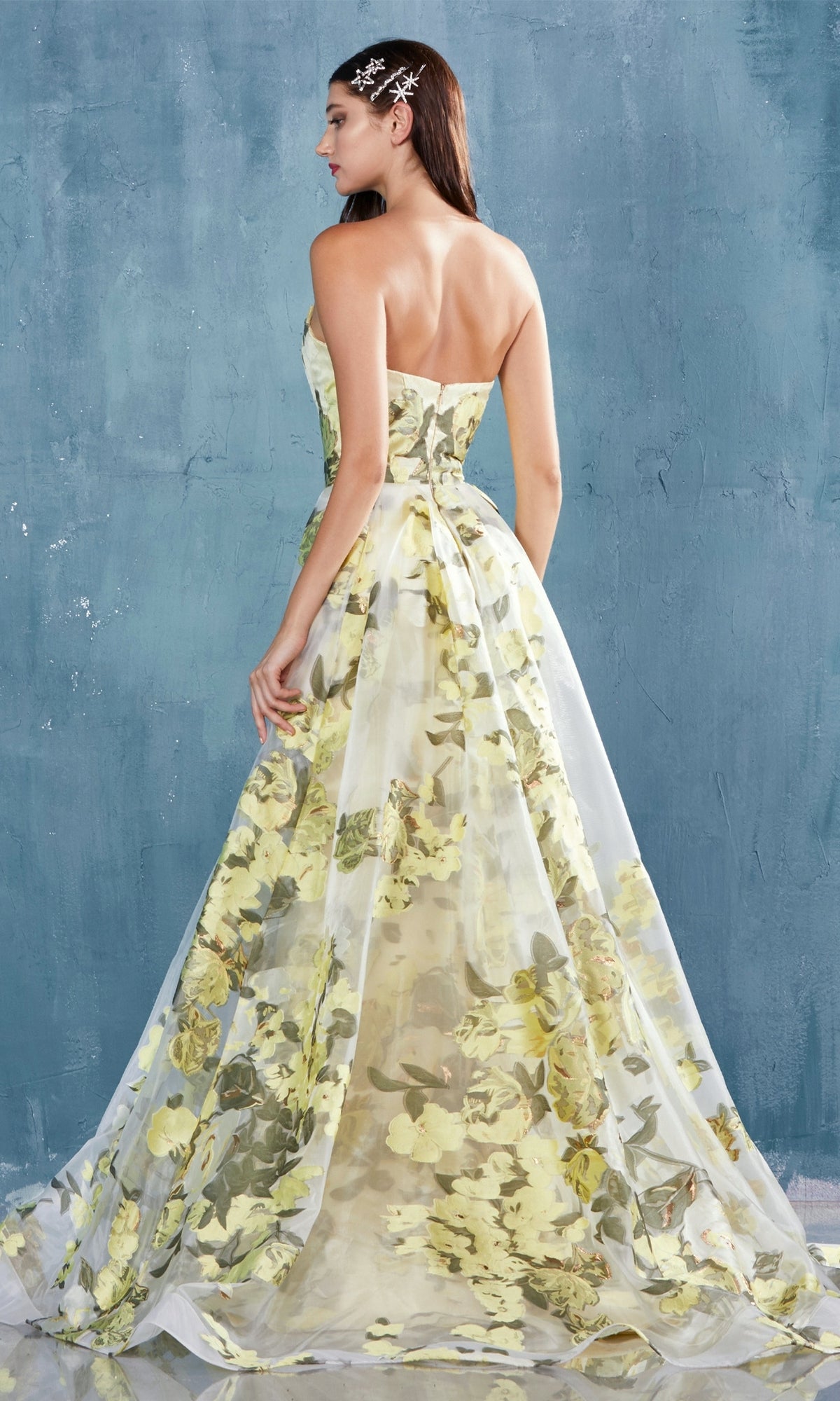 A woman stands facing away, wearing the Lemon Yellow Floral-Print Long Prom Dress A0770 with a strapless design and sheer floral overlay, set against a textured blue background.